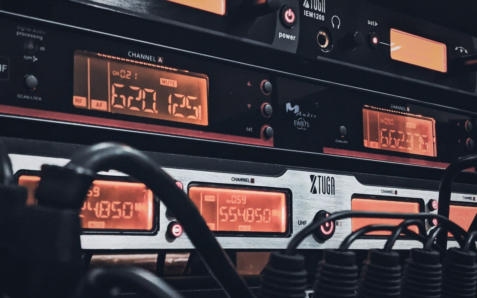 Rack-mounted wireless receivers display glowing orange channel frequencies amid tangled audio cables