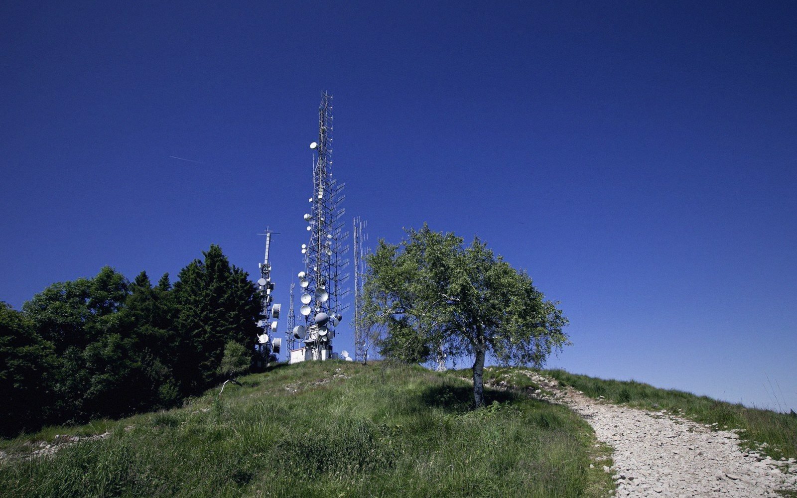 Telecommunications towers with satellite dishes stand atop a grassy hilltop under clear sky