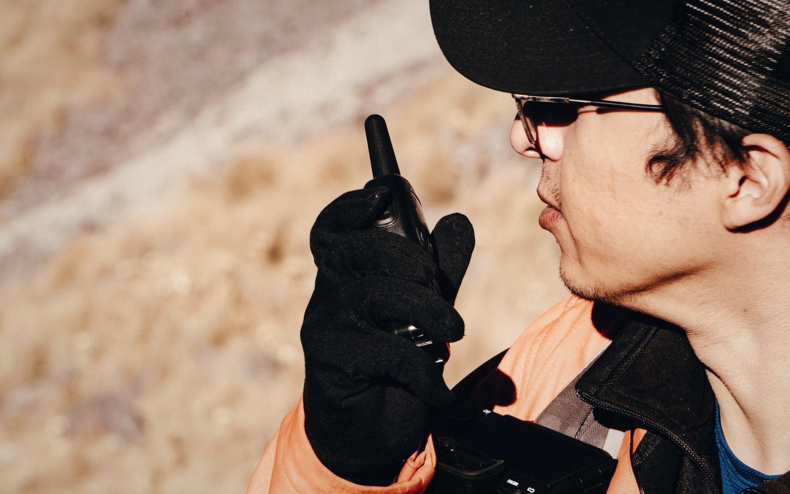 Man in cap and safety vest speaking into a handheld radio outdoors