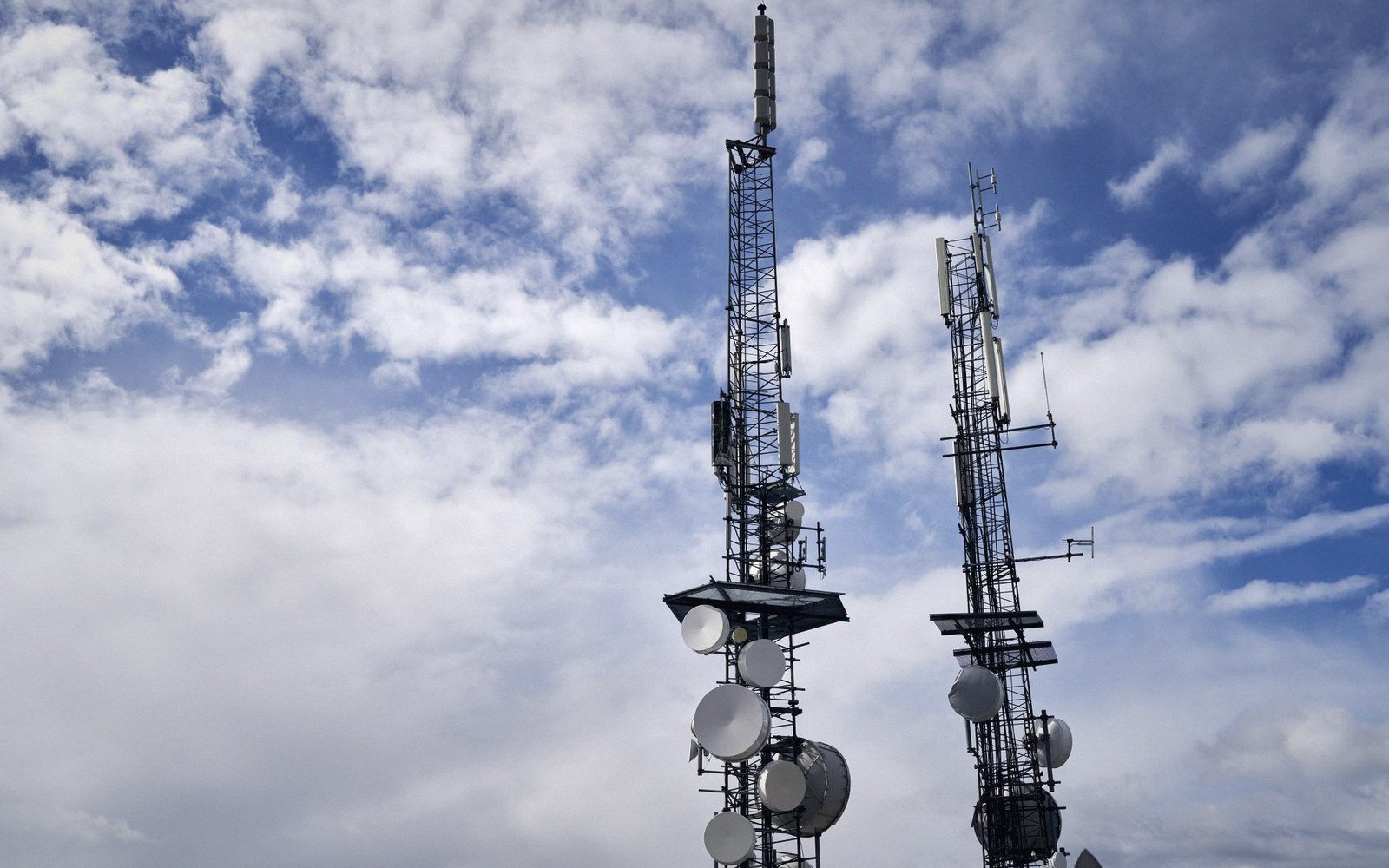 Two telecommunications towers with satellite dishes and antennas against a cloudy sky
