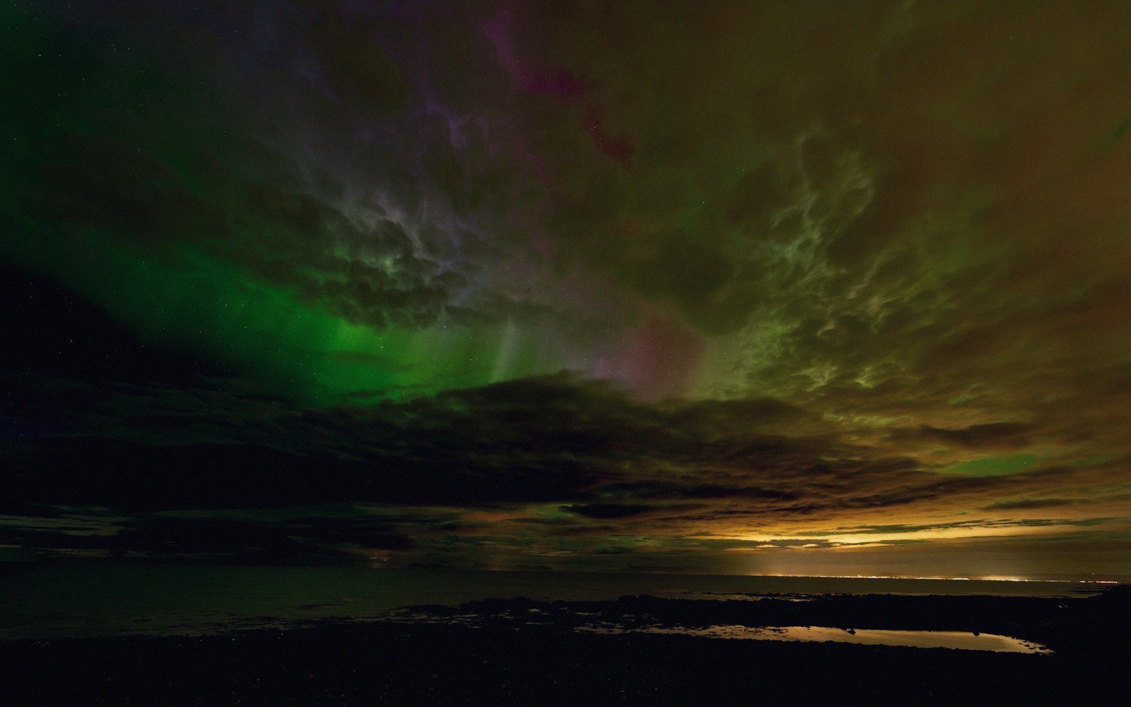 Green and purple aurora glows through clouds above a dark shoreline at dusk